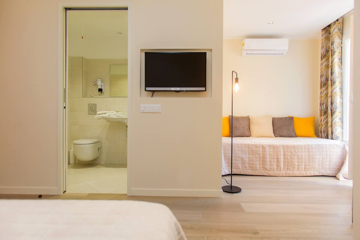 A hotel room with a bed, a television mounted on the wall, a bedside table, and a bathroom visible through an open door.