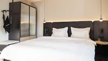 Una habitación de hotel moderna con una cama grande, un armario, una mesita de noche con lámpara y una pequeña mesa redonda con un vaso de agua.