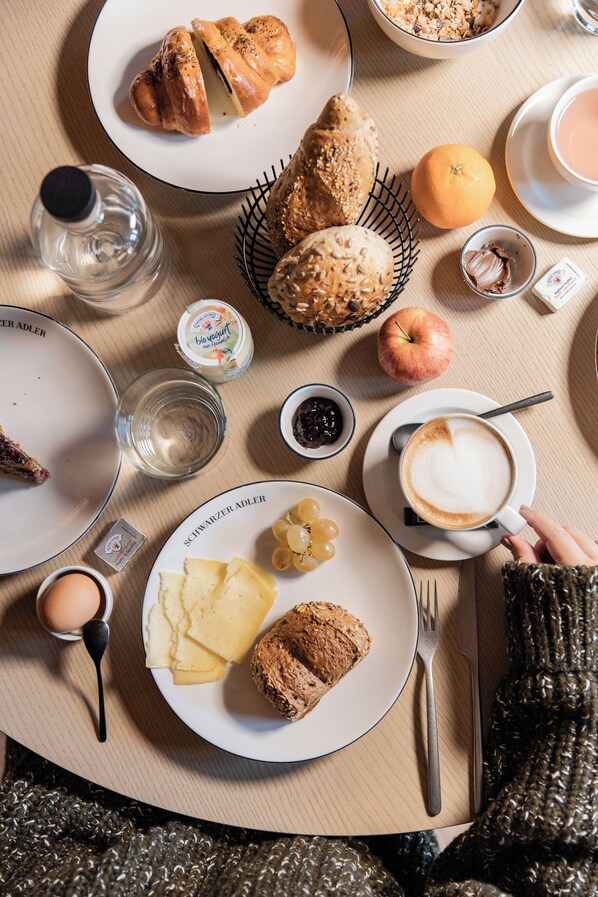 A breakfast table with croissants, an orange, a glass of water, a cup of coffee, and a plate with cheese and grapes. - Schwarzer Adler - Concept Living (Vipiteno)