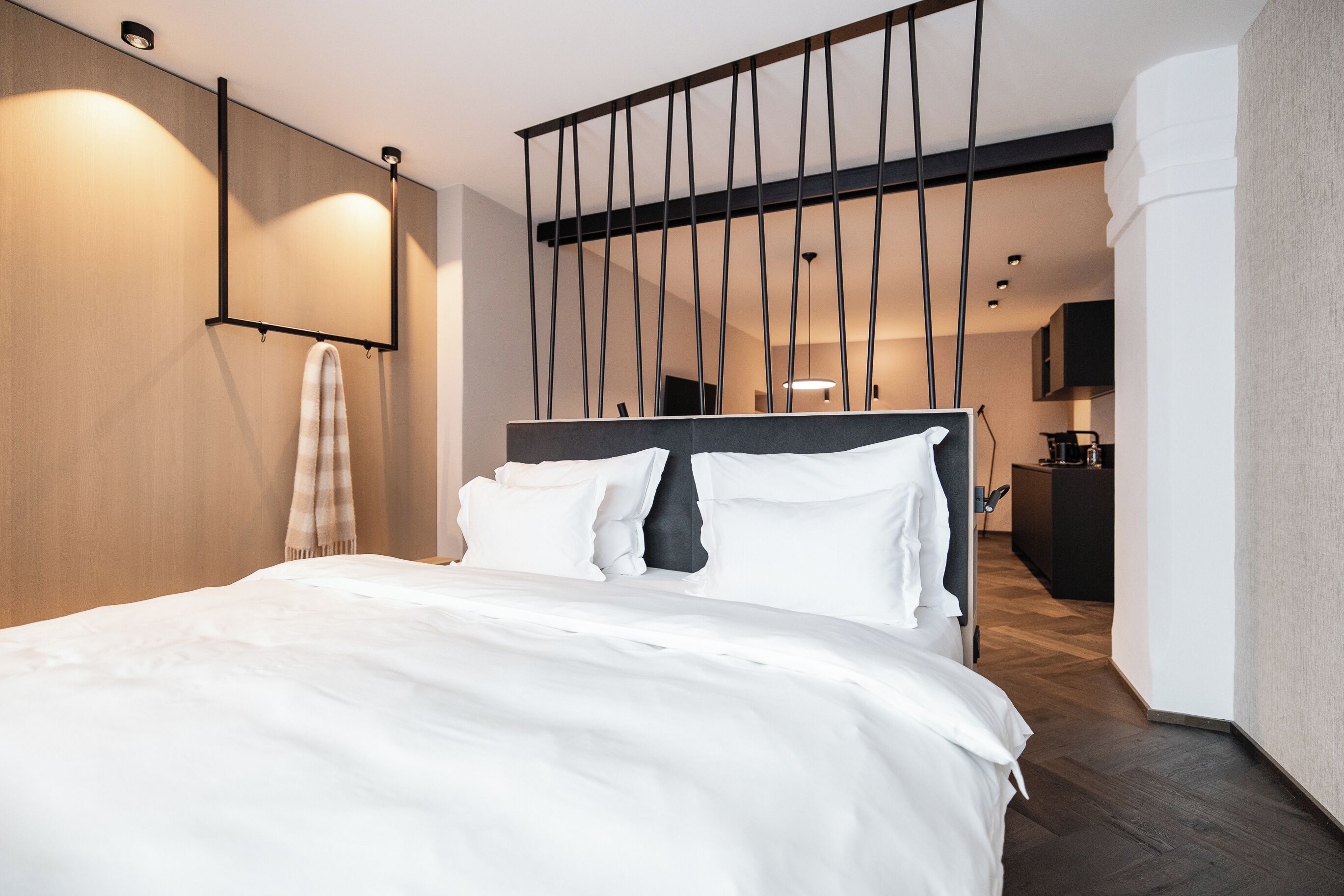 Ein modernes Hotelzimmer mit einem großen Bett, einem Holzkopfbrett, einem aufgehängten Handtuch und Blick in den Küchenbereich.