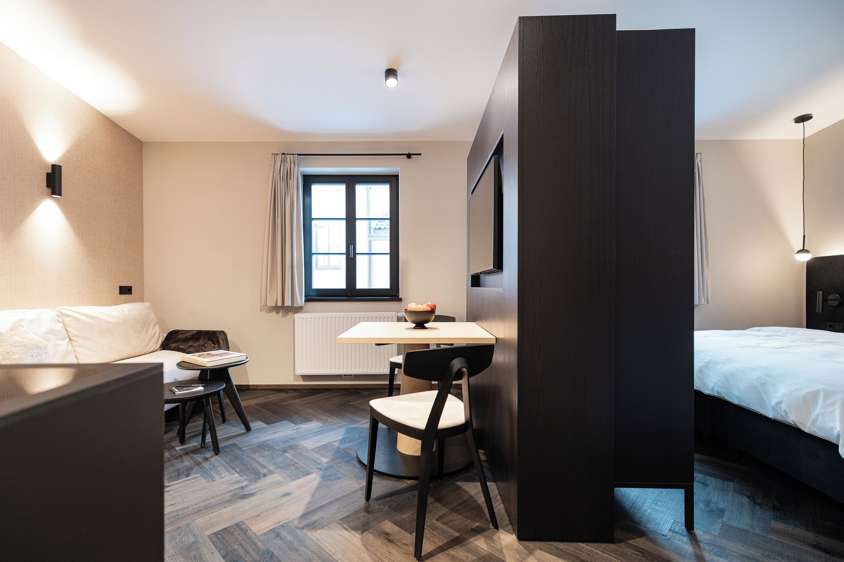 A modern hotel room with a bed, a desk, and a chair. There is a window with curtains, a radiator, and a wall-mounted light fixture.