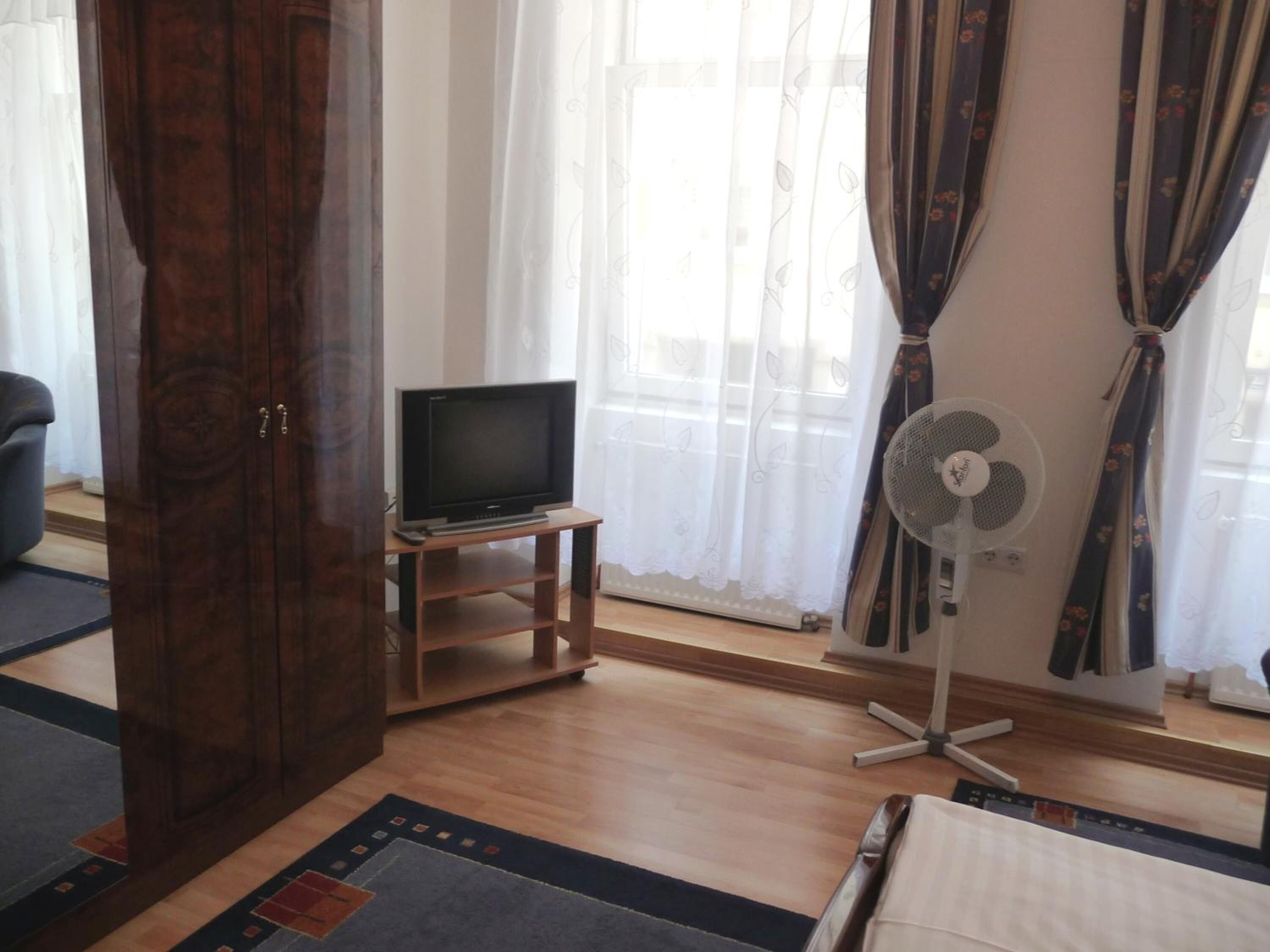 A room with a wooden TV stand, a flat-screen TV, a standing fan, and a bed.
