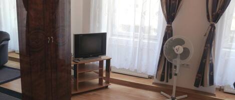A room with a wooden TV stand, a flat-screen TV, a standing fan, and a bed.