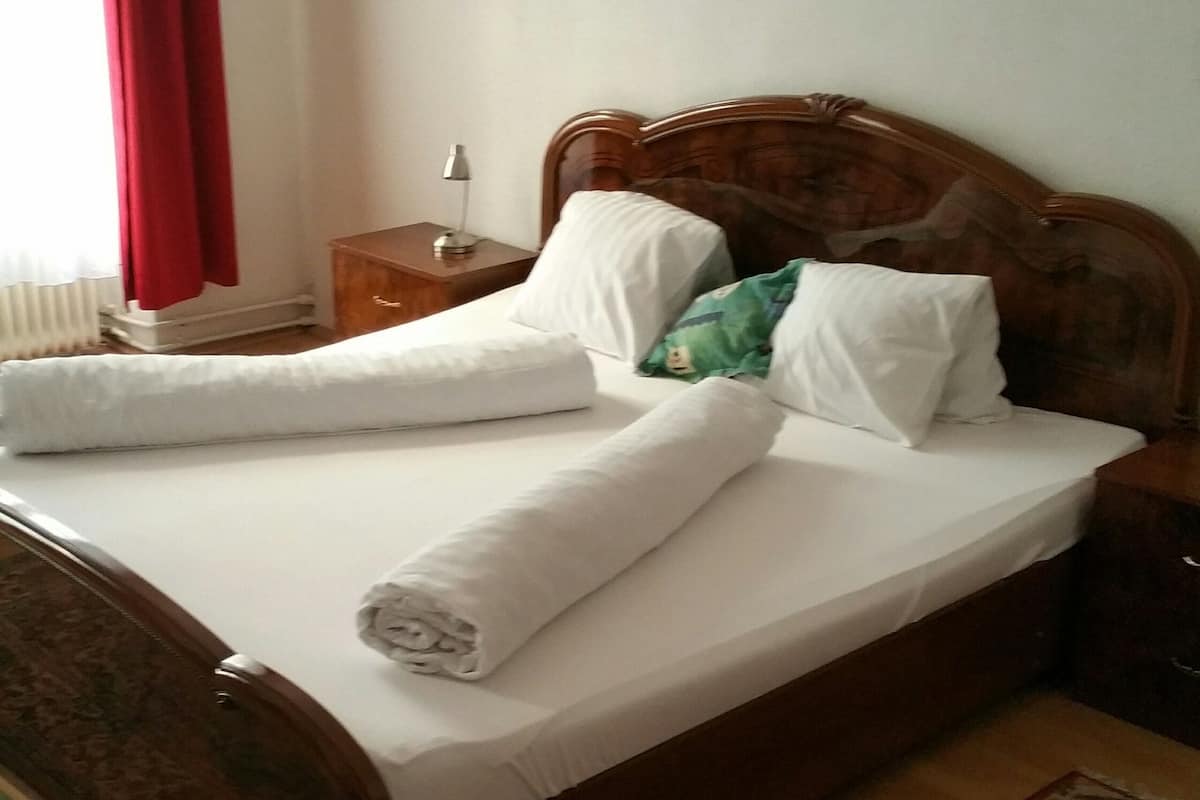Ein ordentlich bezogenes Bett mit weißen Leinentüchern und einem aufgerollten Handtuch, ein hölzernes Kopfteil, ein Nachttisch mit Lampe, ein Bild an der Wand und ein gemustertes Teppich.