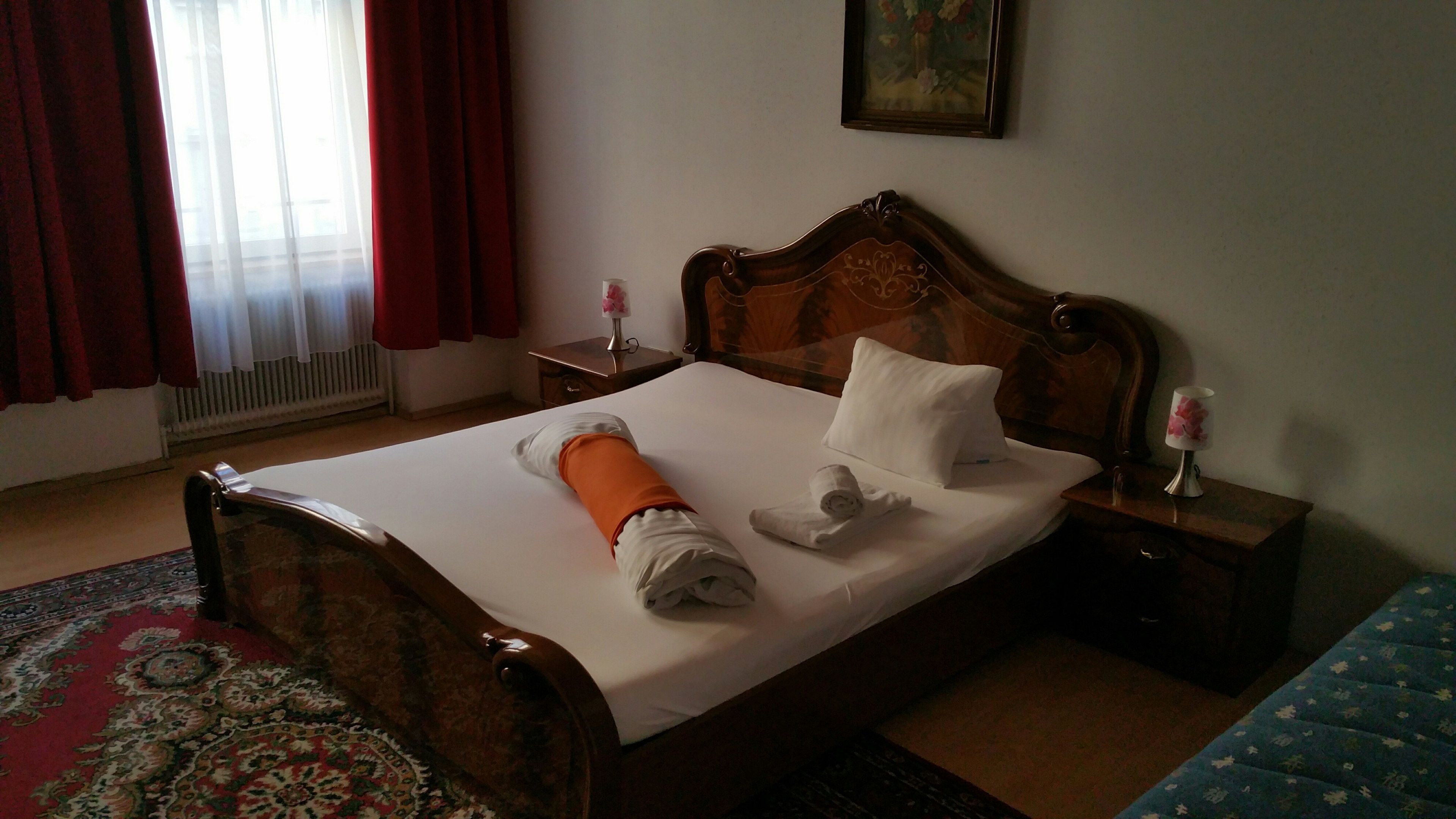 Ein Hotelzimmer mit einem hölzernen Bett, weißen Laken, einem roten und weißen Vorhang, einem kleinen Tisch mit einer Lampe und einem Gemälde an der Wand.