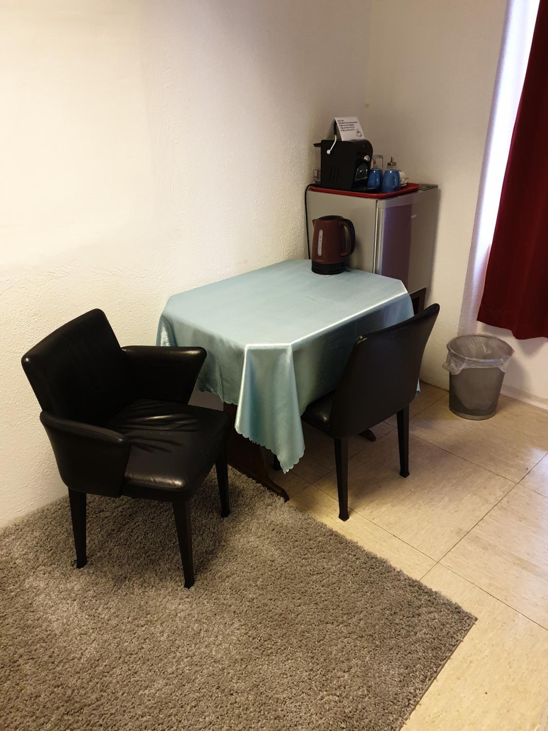 A small dining area with a table covered by a light blue tablecloth, two black chairs, a coffee maker, and a trash can.