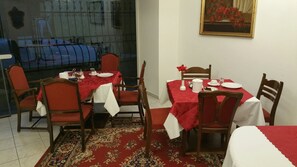 A dining area with two tables set for four, each with a red tablecloth and white napkins. There are four wooden chairs with red cushions. A patterned rug covers the floor. - Appartementhotel Marien-Hof (Vienna)