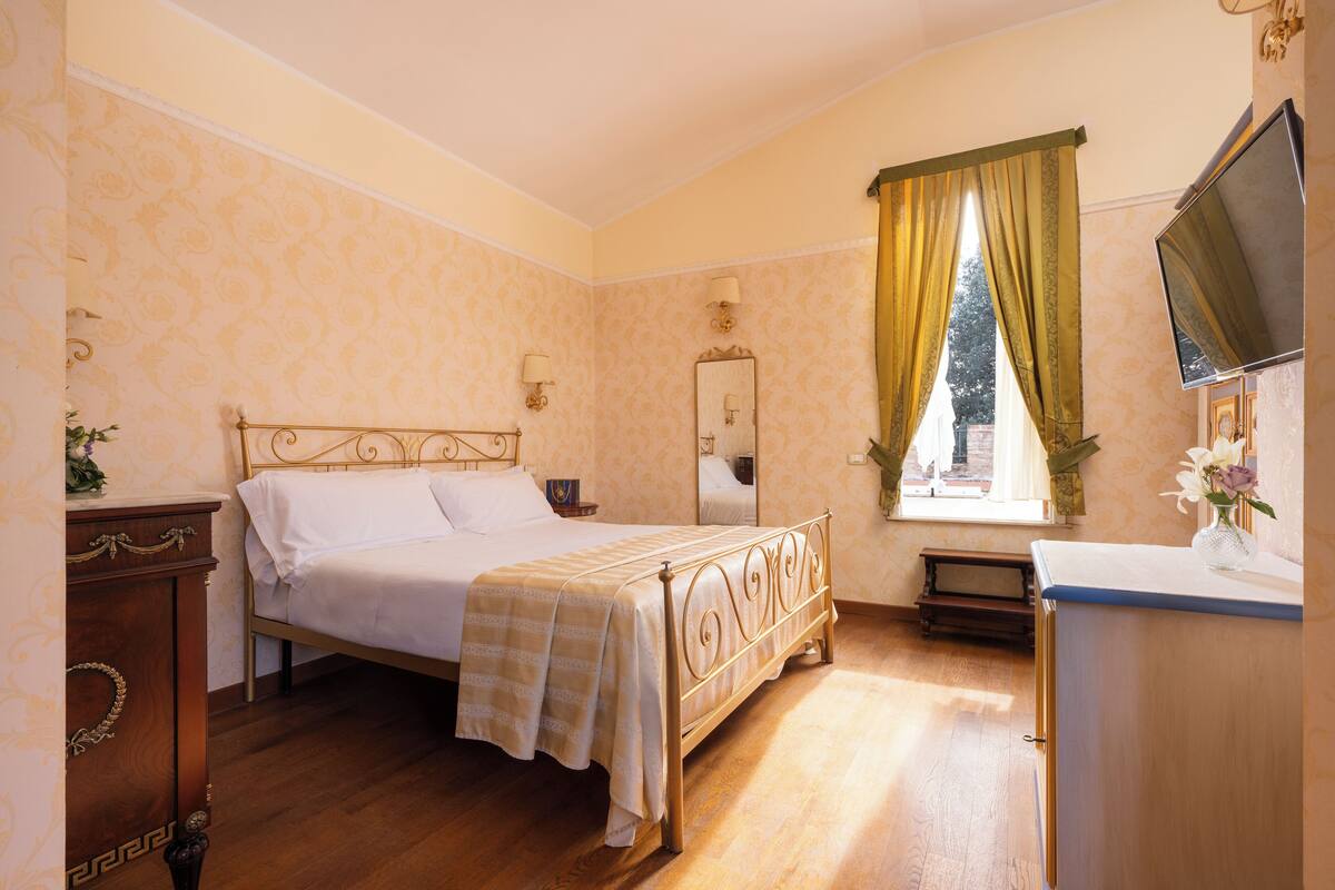 Une chambre à coucher avec un lit, une commode, un miroir, un téléviseur et une fenêtre avec des rideaux.
