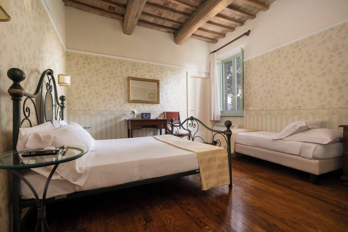 Une chambre avec un plancher en bois, un lit avec un cadre en métal, une table de chevet avec une lampe, un petit bureau et une fenêtre avec des rideaux.