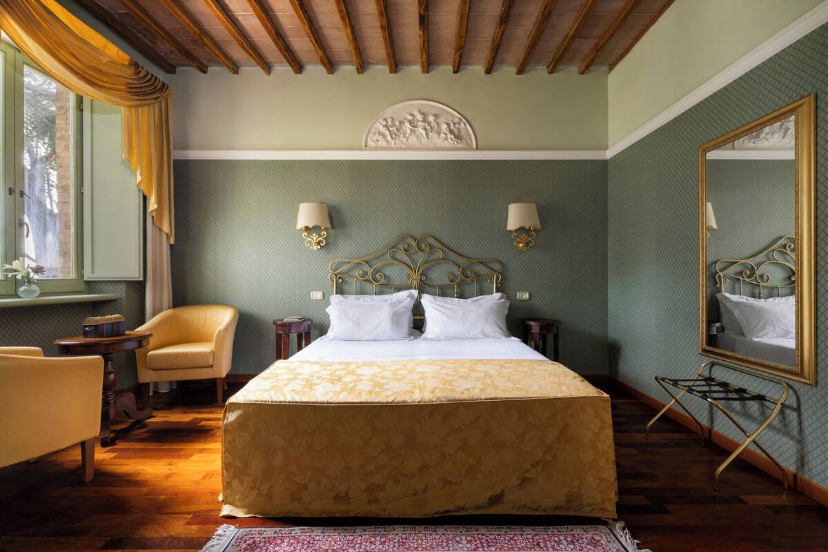 Une chambre à coucher avec un grand lit, deux lampes de chevet, une chaise, une petite table, un miroir et une fenêtre avec des rideaux.