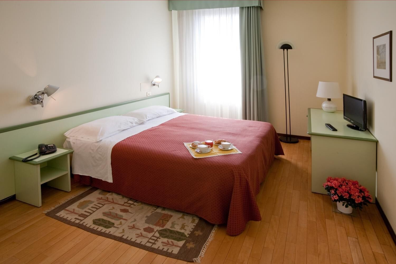 Habitación de hotel con cama, mesita de noche con teléfono, lámpara de noche, televisión, una pequeña mesa con un florero y una ventana con cortinas.