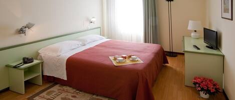 Habitación de hotel con cama, mesita de noche con teléfono, lámpara de noche, televisión, una pequeña mesa con un florero y una ventana con cortinas.