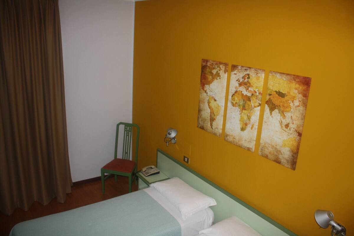 A hotel room with a bed, a chair, a nightstand with a lamp, and a wall with a map.