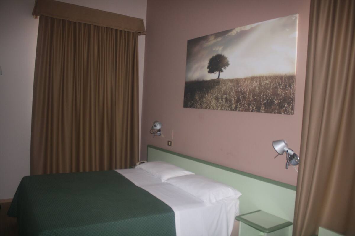 A hotel room with a bed, a nightstand, a lamp, a painting of a tree, and curtains.