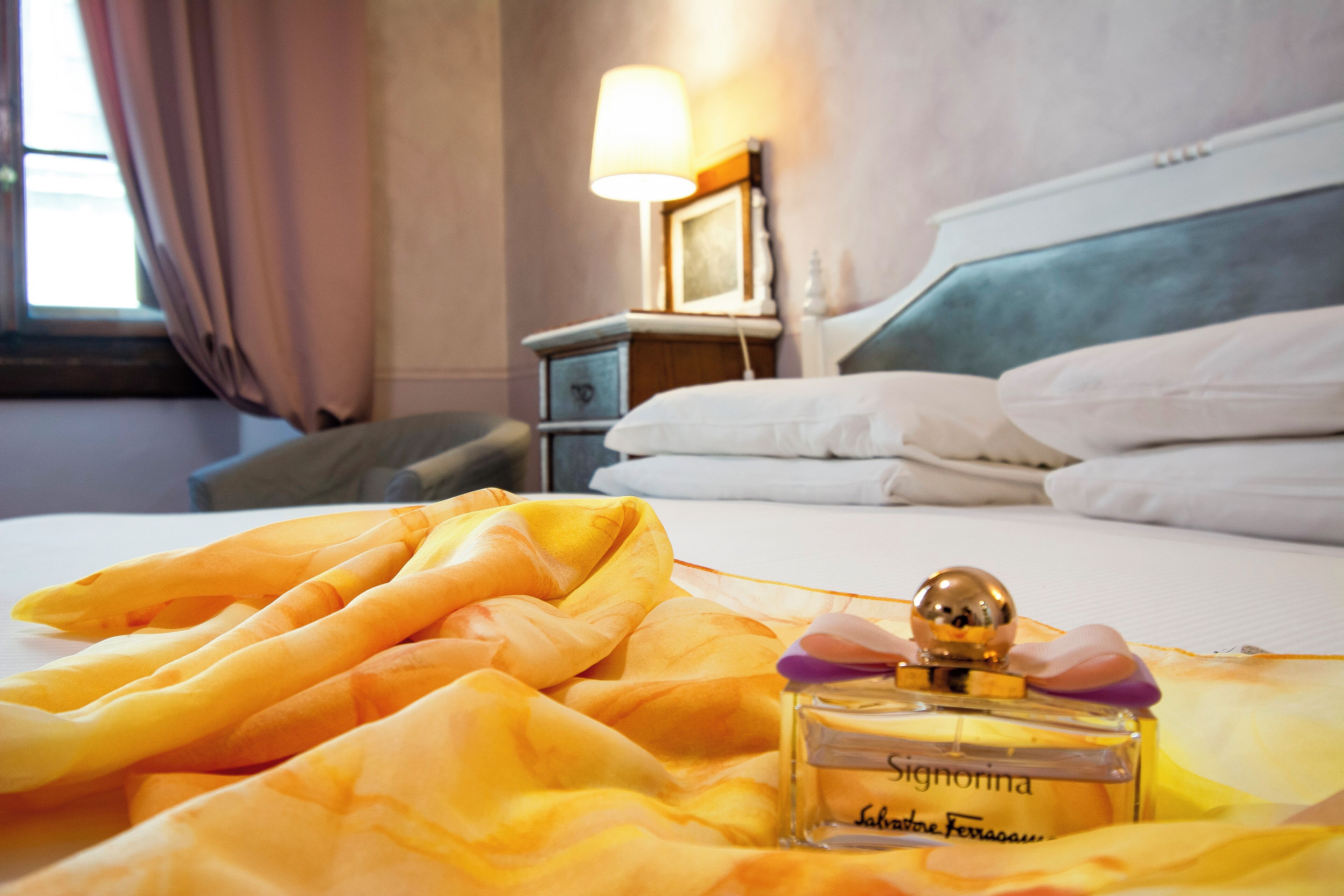 A hotel room with a bed, a bedside lamp, and a bottle of perfume on the bed.