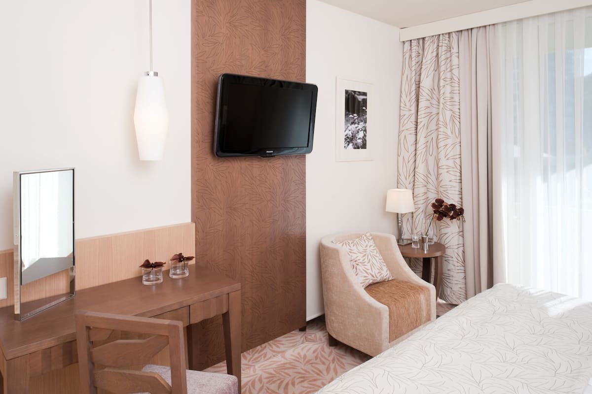 Une chambre d’hôtel dotée d’un téléviseur à écran plat fixé au mur, d’un bureau en bois agrémenté de deux petits vases, d’un fauteuil confortable, d’une table de chevet avec une lampe et d’un lit recouvert d’une couvre-lit à motifs.