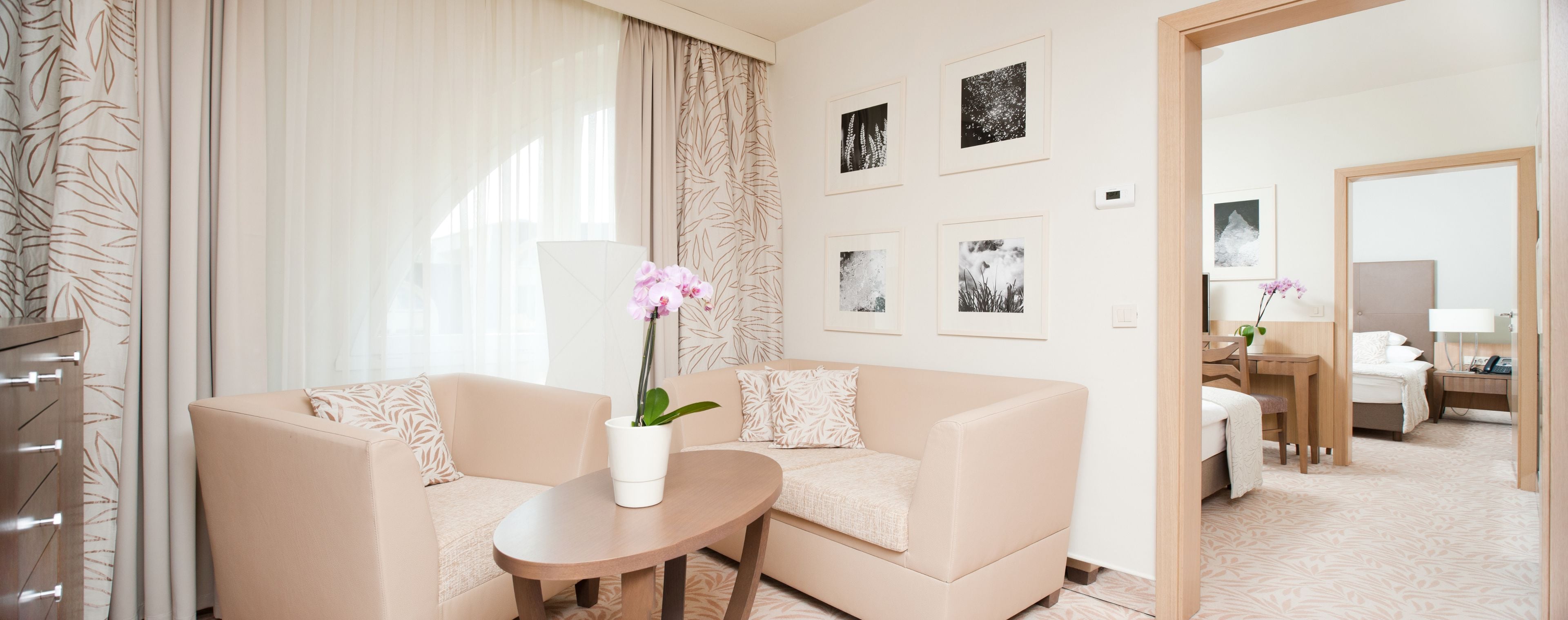 A hotel room with a beige sofa, a small round table, and a potted orchid. There are framed pictures on the wall and a bed in the background.