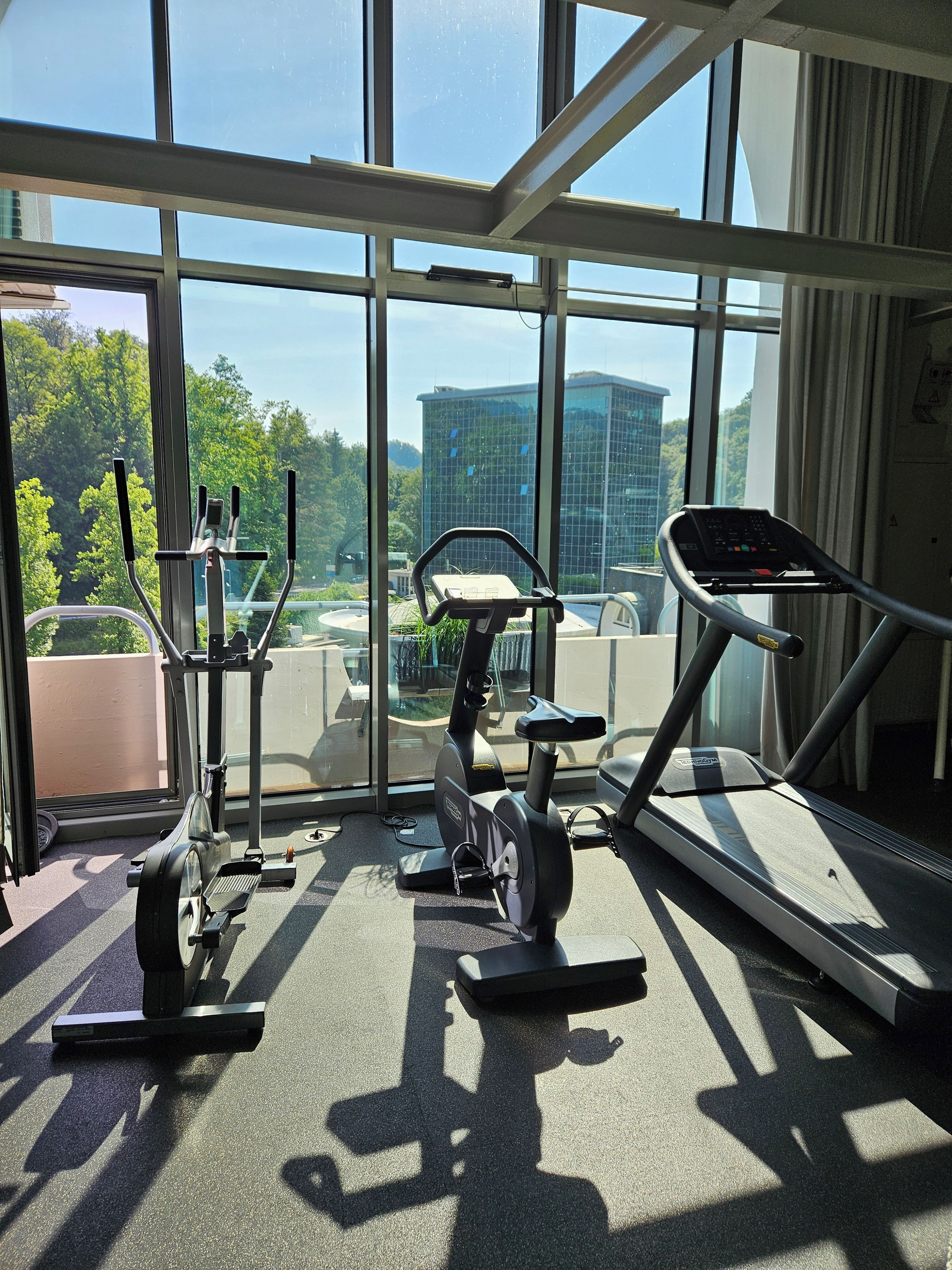 Un gimnasio con equipo de ejercicio y amplias ventanas que ofrecen vistas a un paisaje urbano.