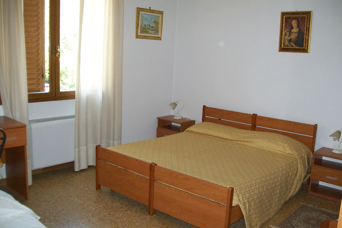 A bedroom with a wooden bed, a bedside table, a nightstand, a window with curtains, and two framed pictures on the wall.