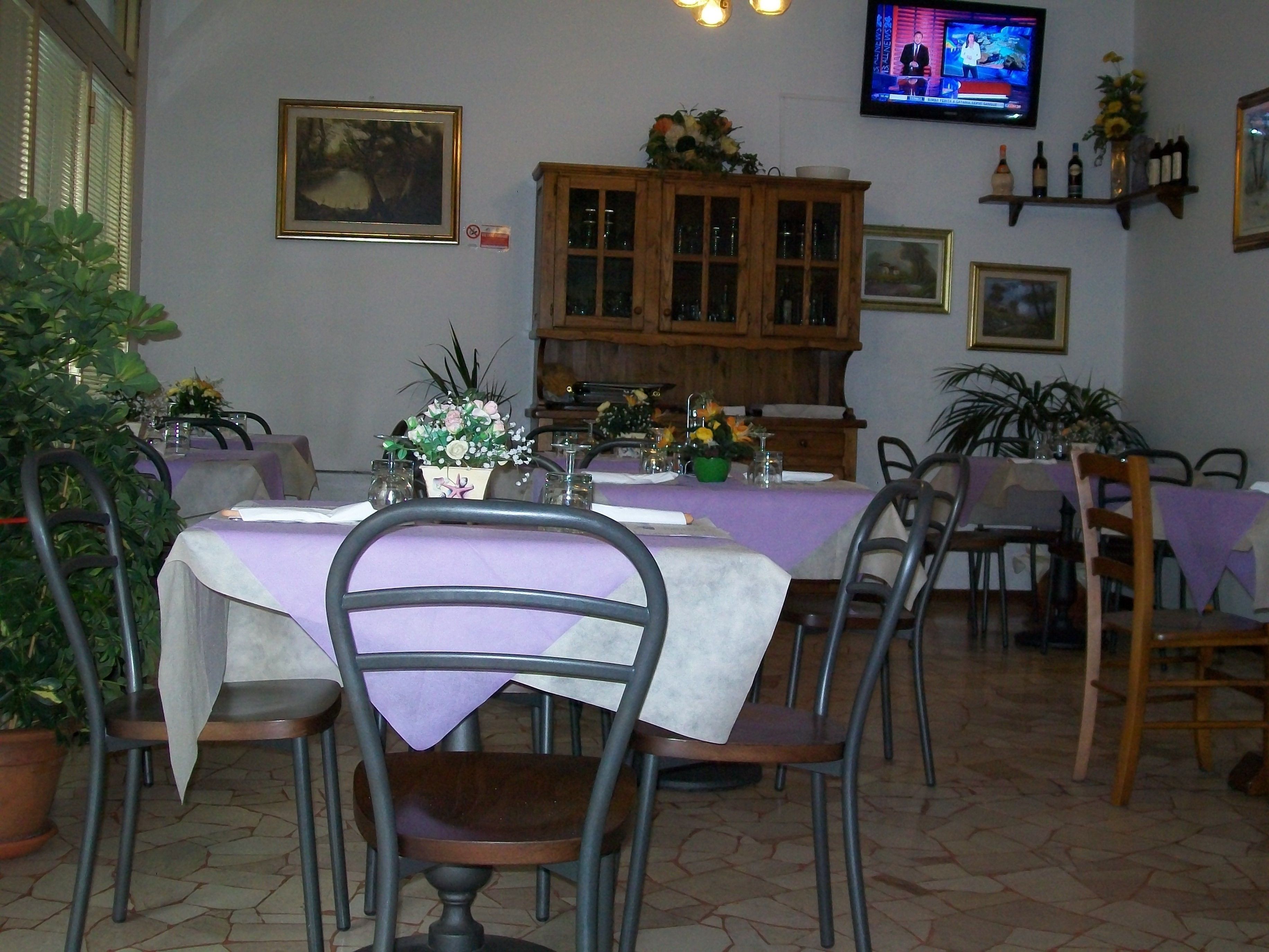 A dining area with tables set for four, chairs, a wooden cabinet, and a television mounted on the wall.
