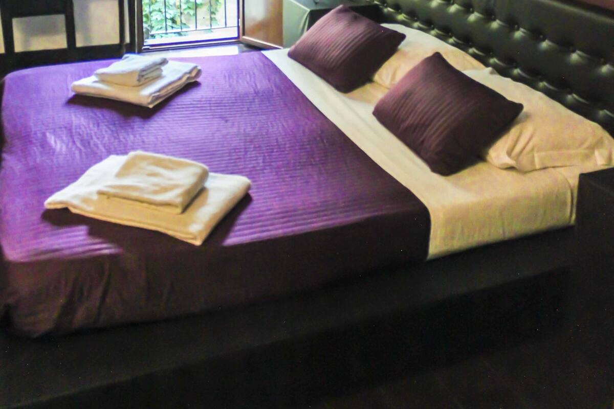 A hotel room with a large bed, a desk, a chair, a balcony with a view, and a painting on the wall.