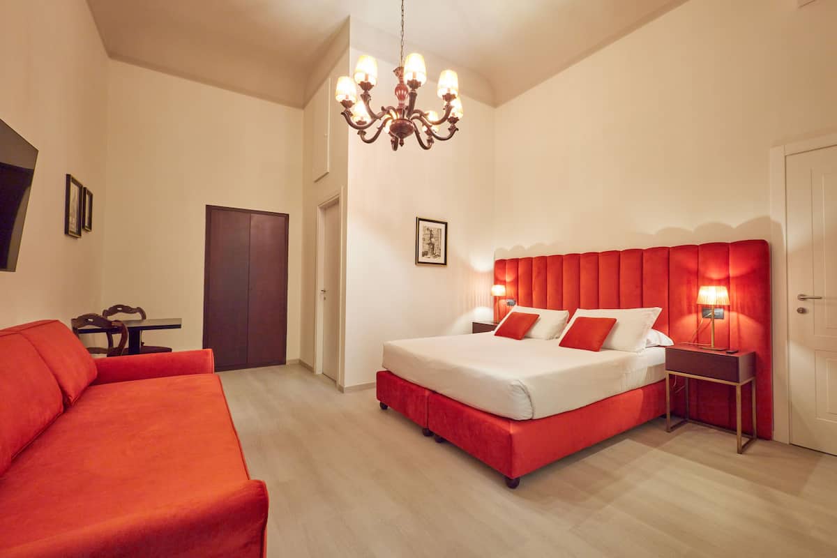 A bedroom with a large bed, a red upholstered sofa, a chandelier, and a small table with a lamp.