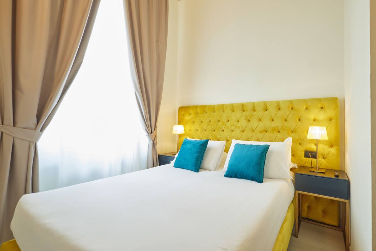 A hotel room with a large bed, two bedside tables, a yellow headboard, and a window with curtains.