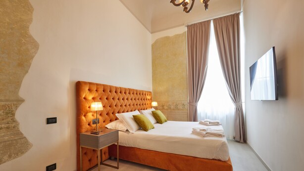 A hotel room with a tufted headboard, a bedside table with a lamp, a neatly made bed with pillows, and a flat-screen TV mounted on the wall. - Residenza Maglioni al Battistero (Florence)
