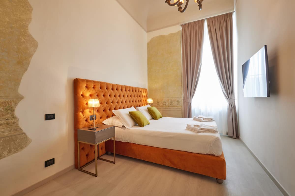 A hotel room with a tufted headboard, a bedside table with a lamp, a neatly made bed with pillows, and a flat-screen TV mounted on the wall.