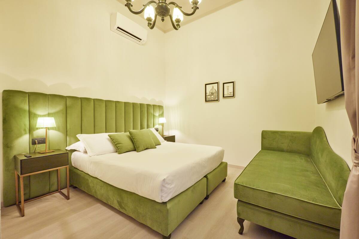 A hotel room with a green upholstered bed, a matching green armchair, a nightstand with a lamp, and a wall-mounted TV.