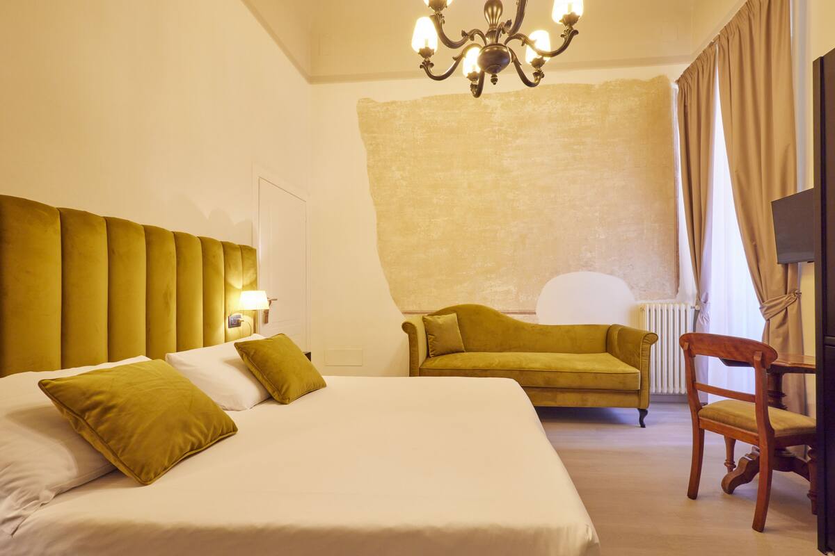 A hotel room with a large bed, a sofa, a wooden chair, a television, and a chandelier.