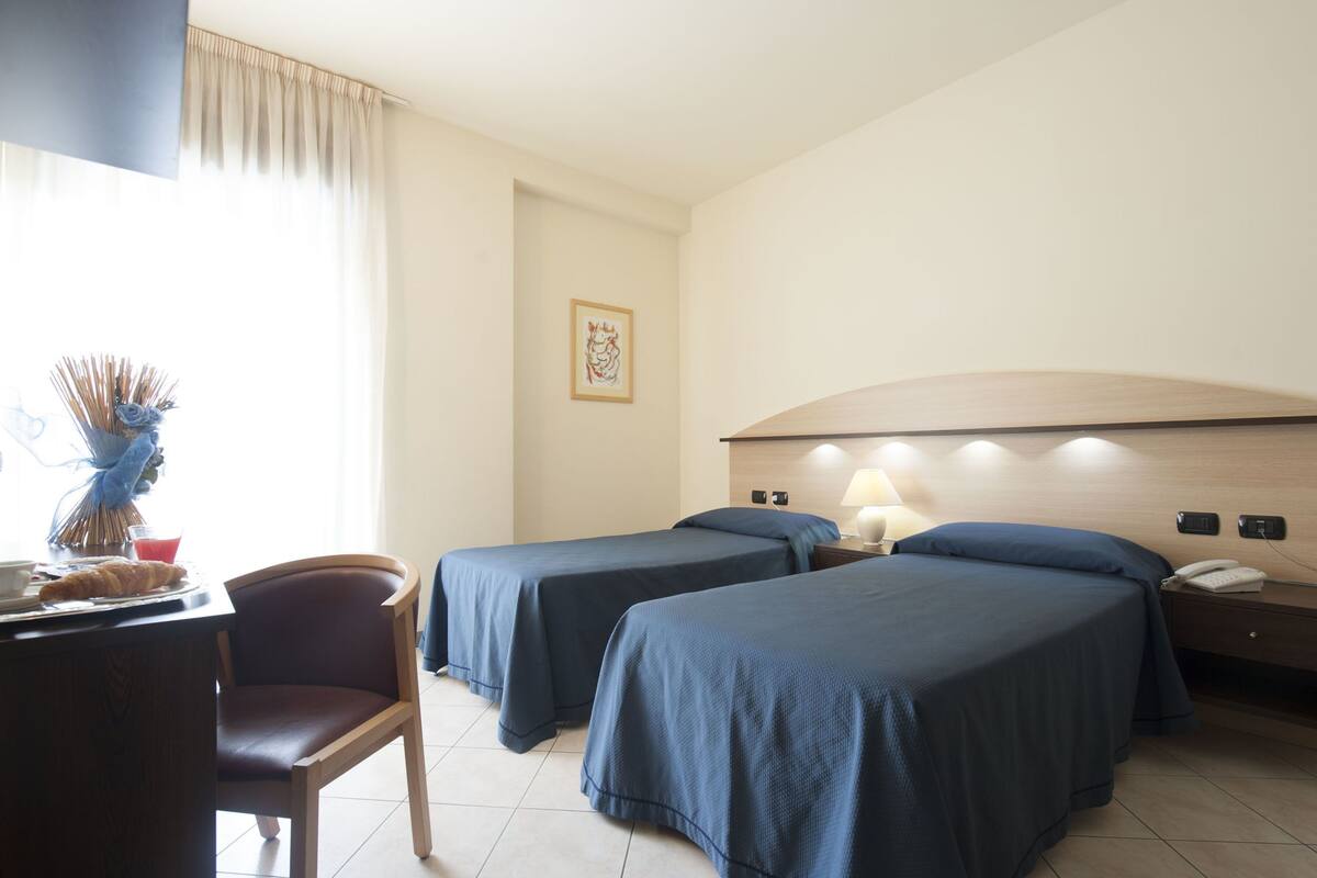Habitación de hotel con dos camas individuales, un escritorio con una silla, una lámpara y una pequeña mesa con un florero y un croissant.