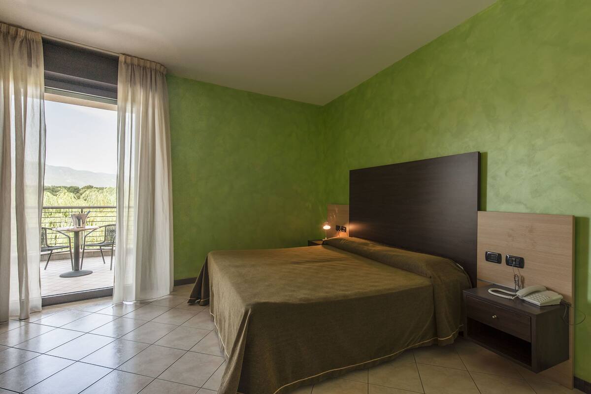 Habitación de hotel con cama, mesita de noche con lámpara, teléfono y vistas a un patio con mesa y sillas.