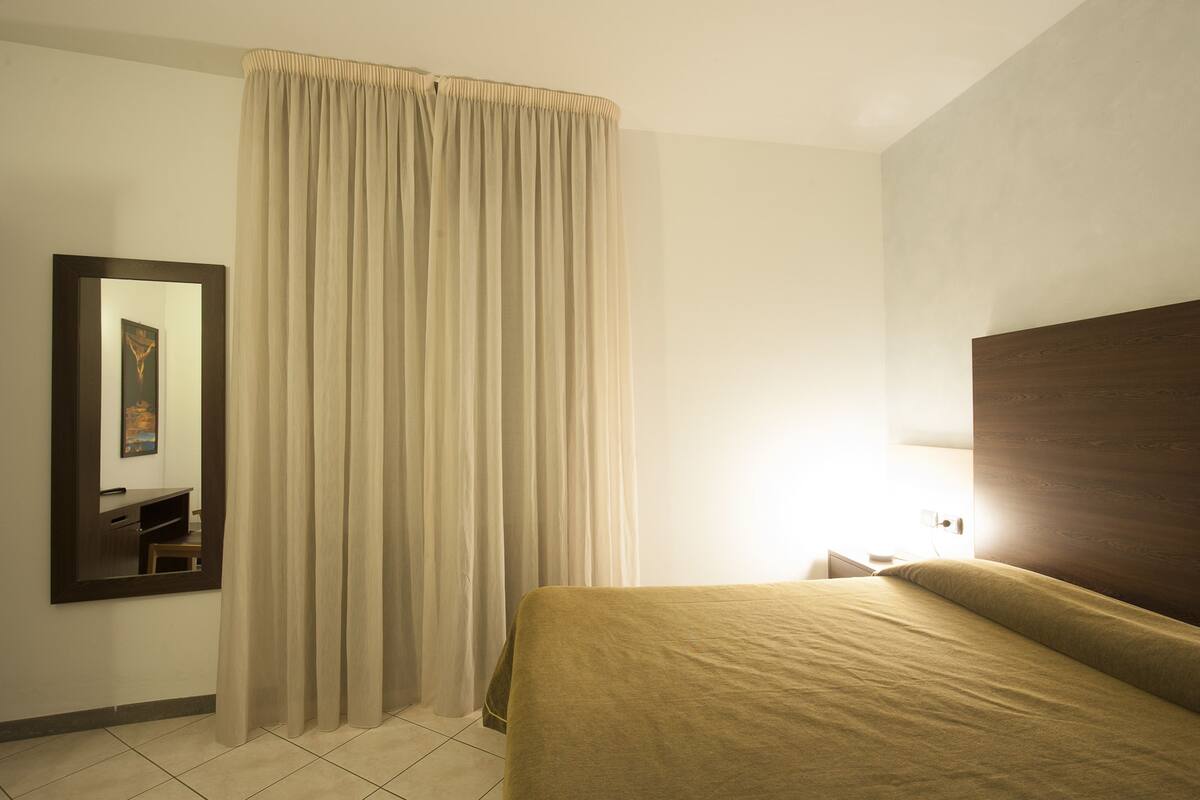 Una habitación de hotel con una cama, una mesita de noche, un espejo y una cortina.