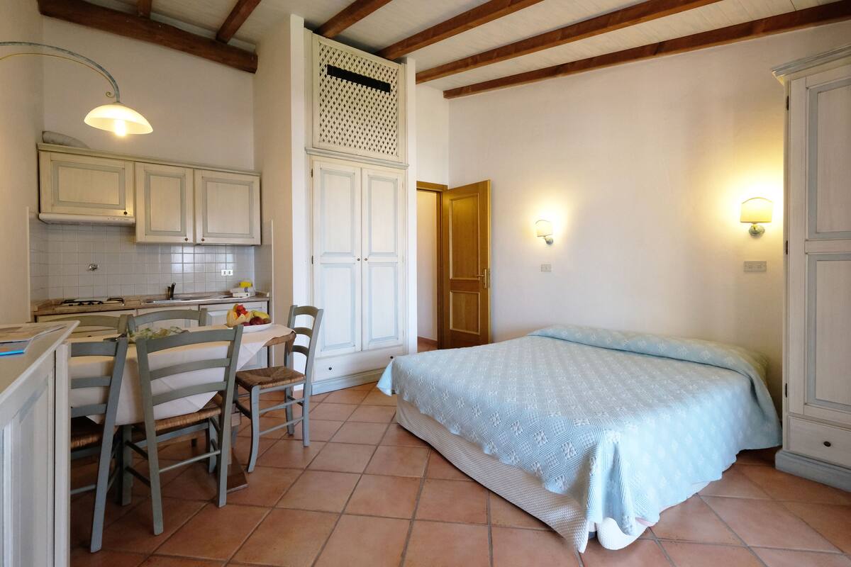 A small, cozy room with a bed, a dining table with chairs, a kitchen area, and a wooden ceiling.