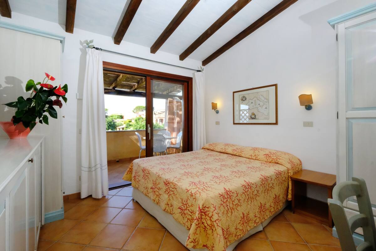 A bedroom with a bed, a potted plant, a desk, a chair, a window with curtains, and a balcony with a table and chairs.
