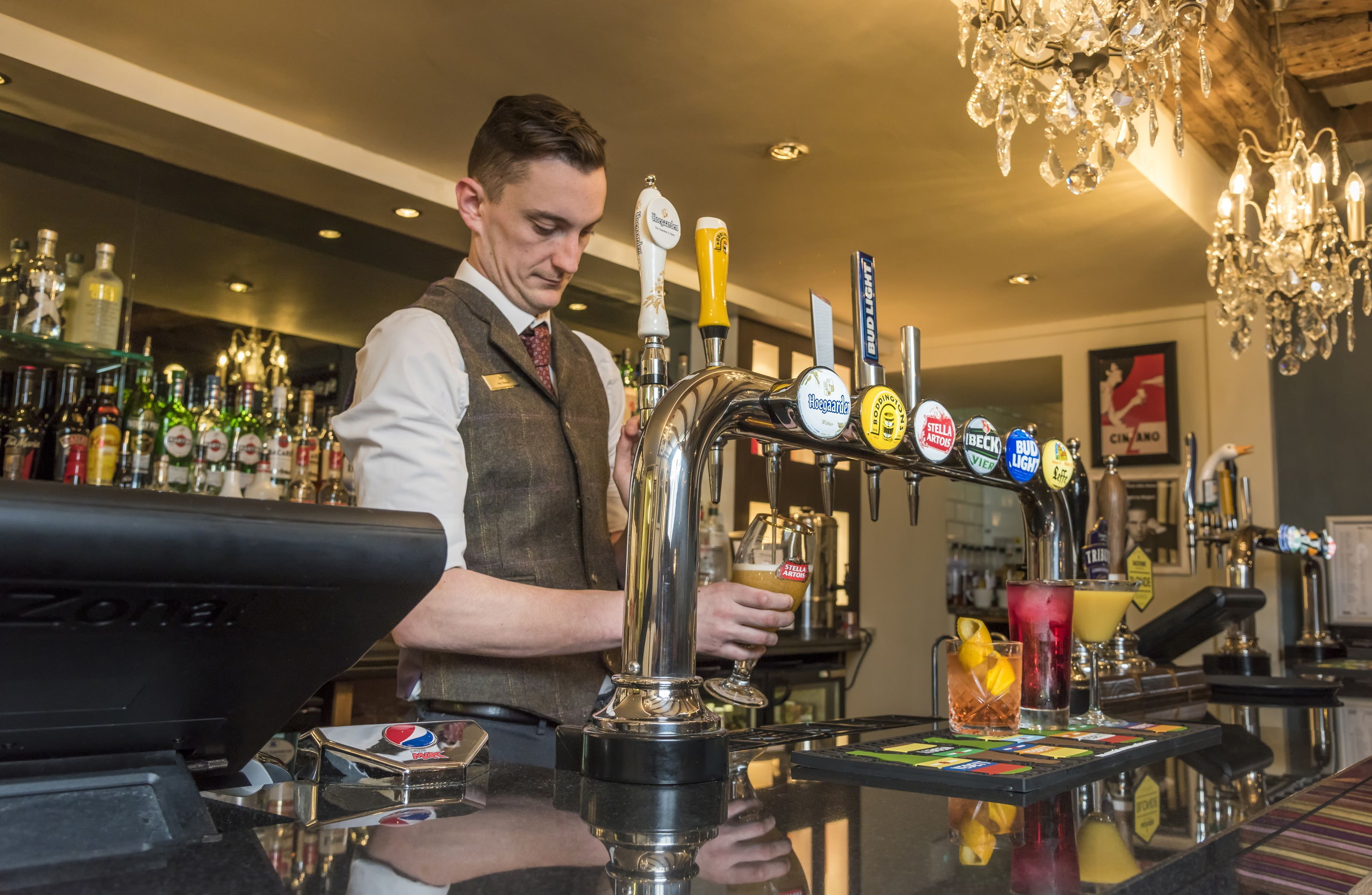 Un barman derrière un comptoir dans un bar doté de plusieurs robinets à bière et où l’on peut voir, en arrière-plan, une variété de bouteilles.