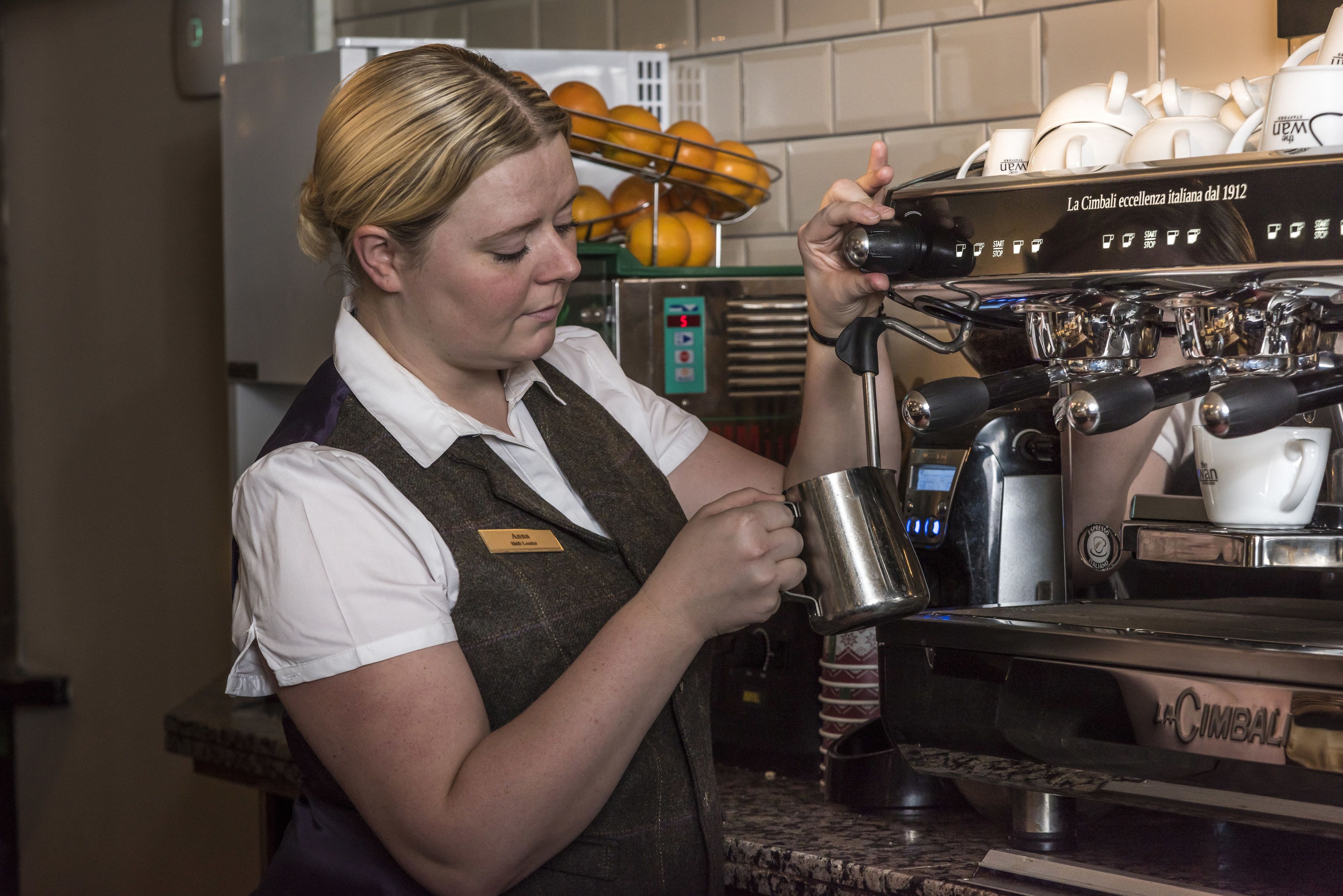 En barista som gör i ordning en dryck vid en kaffemaskin, bärandes ett namnmärke och en uniform.
