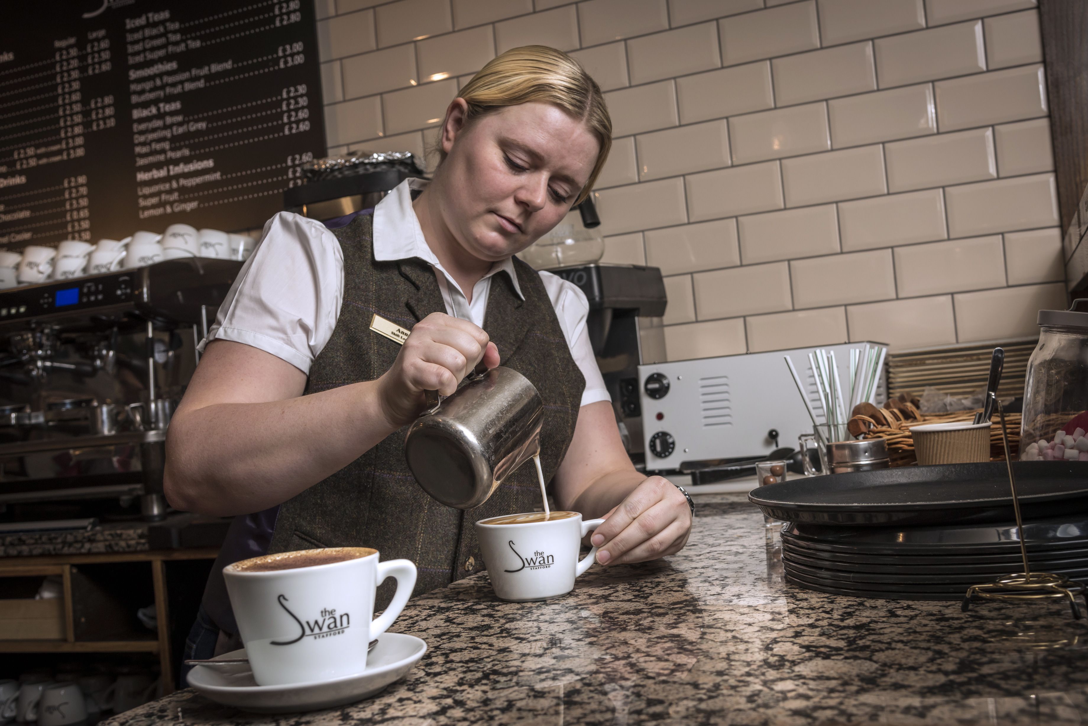 En barista hælder mælk i en kop med teksten 'The Swan' i en café.