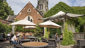 An outdoor patio with wicker tables and umbrellas, a brick building with a tower in the background, and lush greenery.