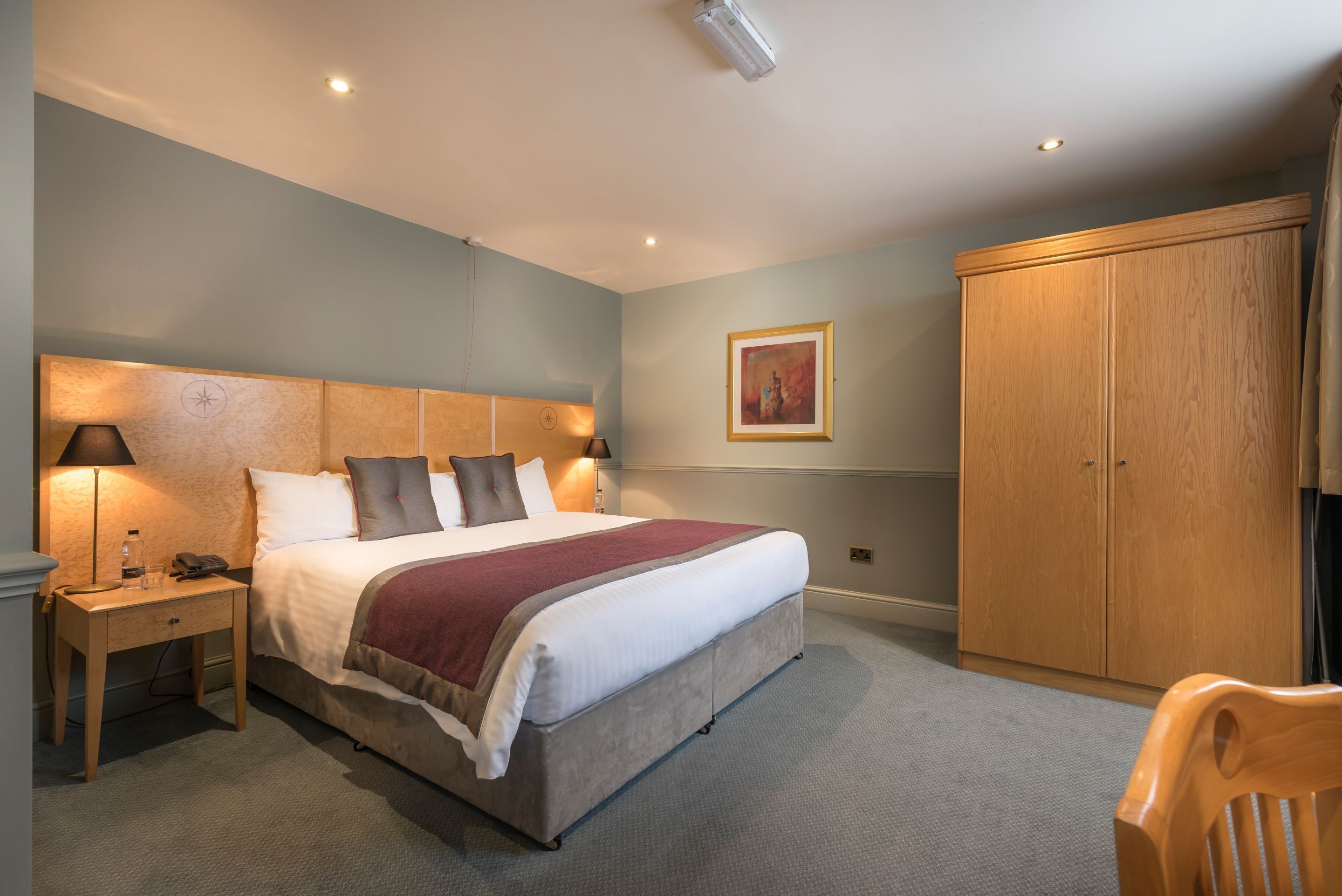 A bedroom with a bed, bedside table, lamp, wardrobe, and a framed picture on the wall.