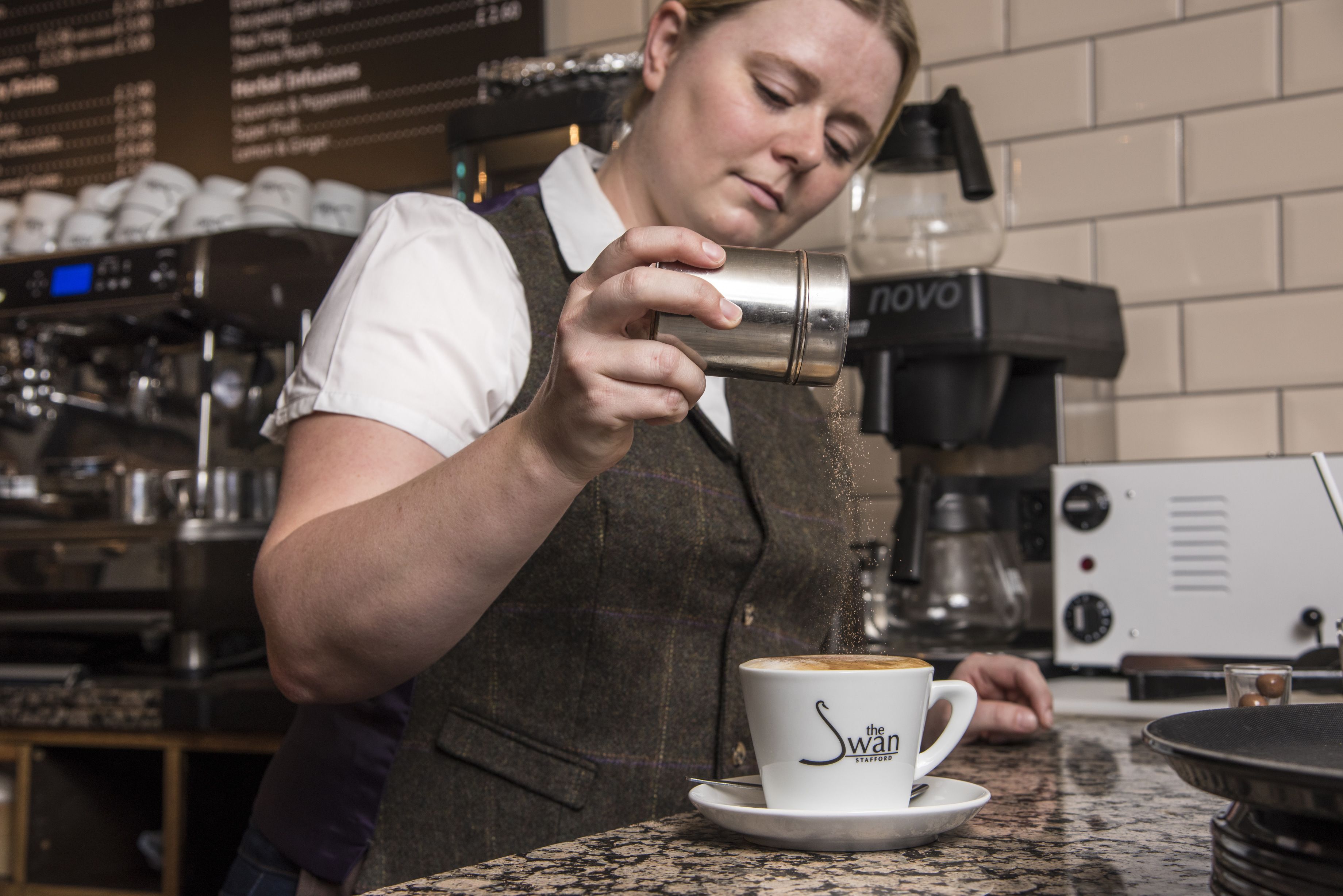 A person in a white shirt and brown vest preparing a cup of coffee with a coffee machine.