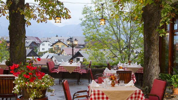 An outdoor dining area with tables set for two, surrounded by trees and a view of buildings in the background. - Gostilna Lectar (Radovljica)