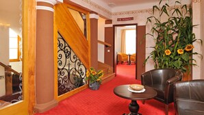A hotel lobby with a red carpet, a staircase with a decorative metal railing, a large potted sunflower plant, leather chairs, and a table with a placemat. - Gostilna Lectar (Radovljica)