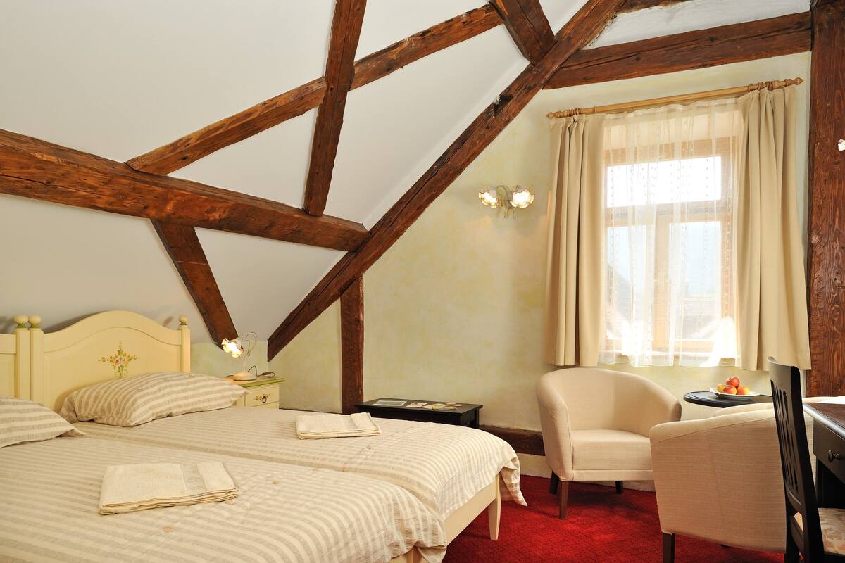 A bedroom with a wooden ceiling, two beds, a chair, a desk, and a window with curtains.