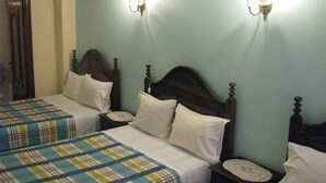 Een hotelkamer met twee eenpersoonsbedden, elk met een houten hoofdbord en witte lakens. Er staan nachtkastjes met lampen en een klein decoratief object op elk. De muren zijn in een lichte kleur geschilderd, en aan de linkerkant van de afbeelding hangt een gordijn.