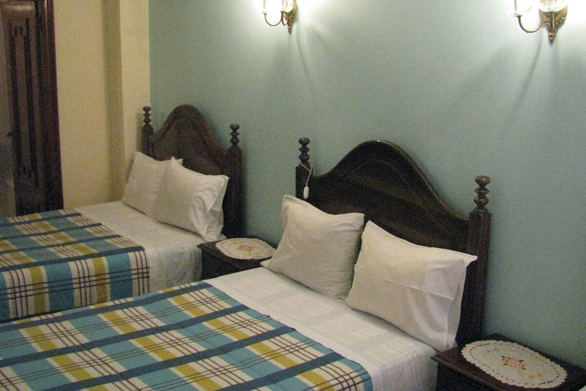 Une chambre d’hôtel avec deux lits simples, chacun doté d’une tête de lit en bois et de linge de lit blanc. On y trouve des tables de chevet avec des lampes et un petit objet décoratif sur chacune. Les murs sont peints dans une couleur claire, et un rideau se trouve sur le côté gauche de l’image.