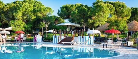 A pool area with lounge chairs and umbrellas, surrounded by trees and a clear sky.