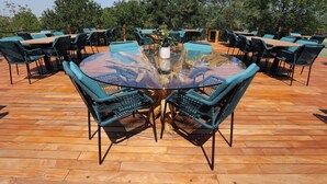 Een terras met ronde glazen tafels omringd door groene stoelen, gelegen op een houten terras met uitzicht op bomen.