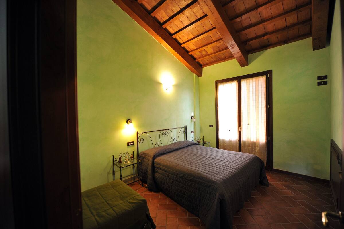 A bedroom with a bed, a nightstand, a lamp, a window with curtains, and a wooden ceiling.